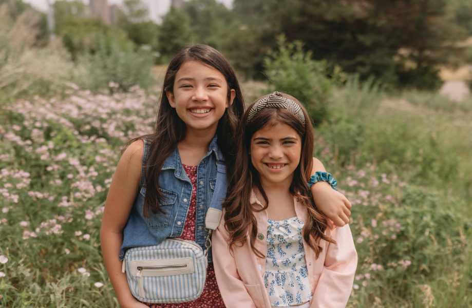 Two young girls standing next to each other in a field with flowers in background.